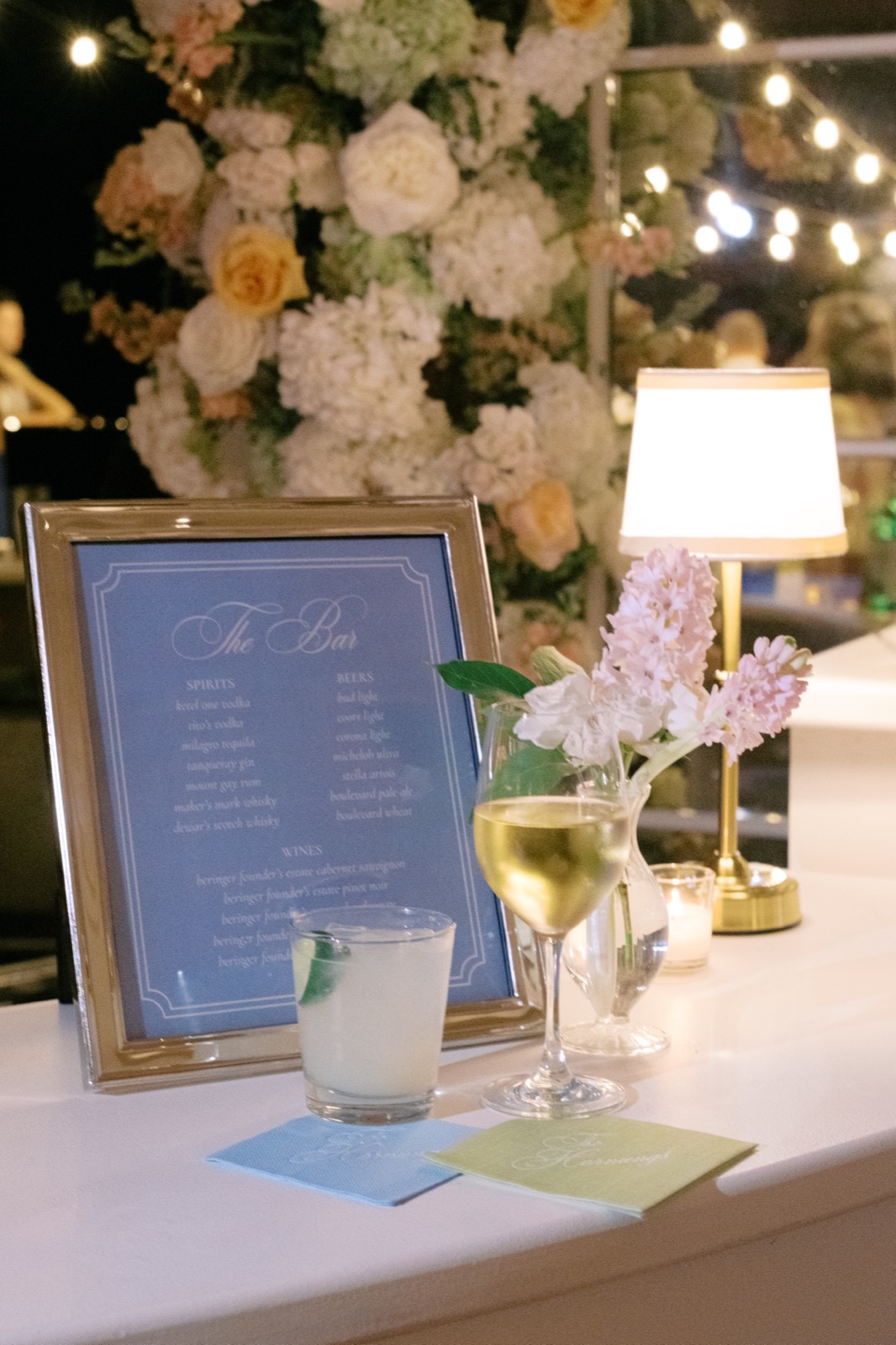 A framed bar menu in soft blue lettered with spirits, beers, and wines, set on a bar with a lush floral arch and bar napkins.