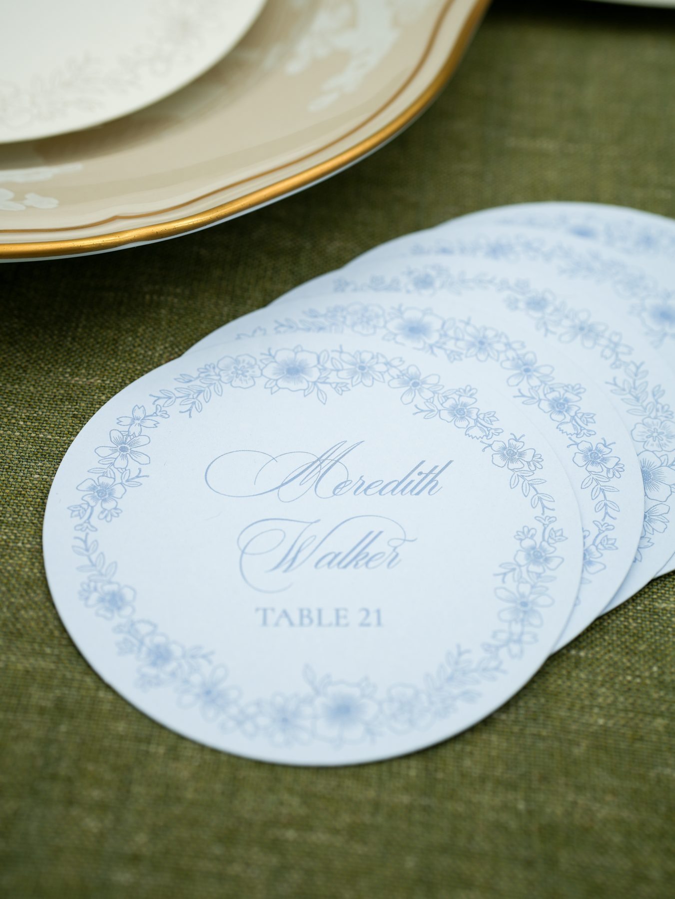 A stack of round blue floral wedding place cards next to a gold-rimmed china plate on olive linen.