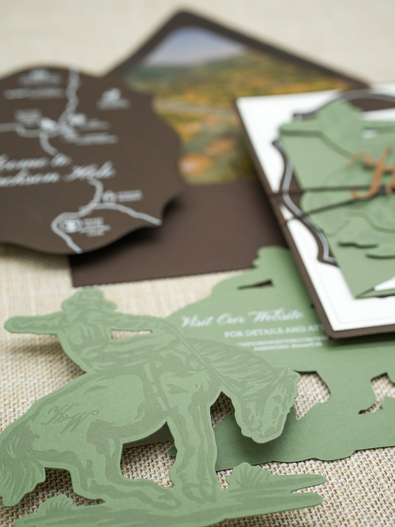 Close-up of a sage green die-cut cowboy and horse silhouette belly band layered over a custom watercolor map for a mountain wedding suite.