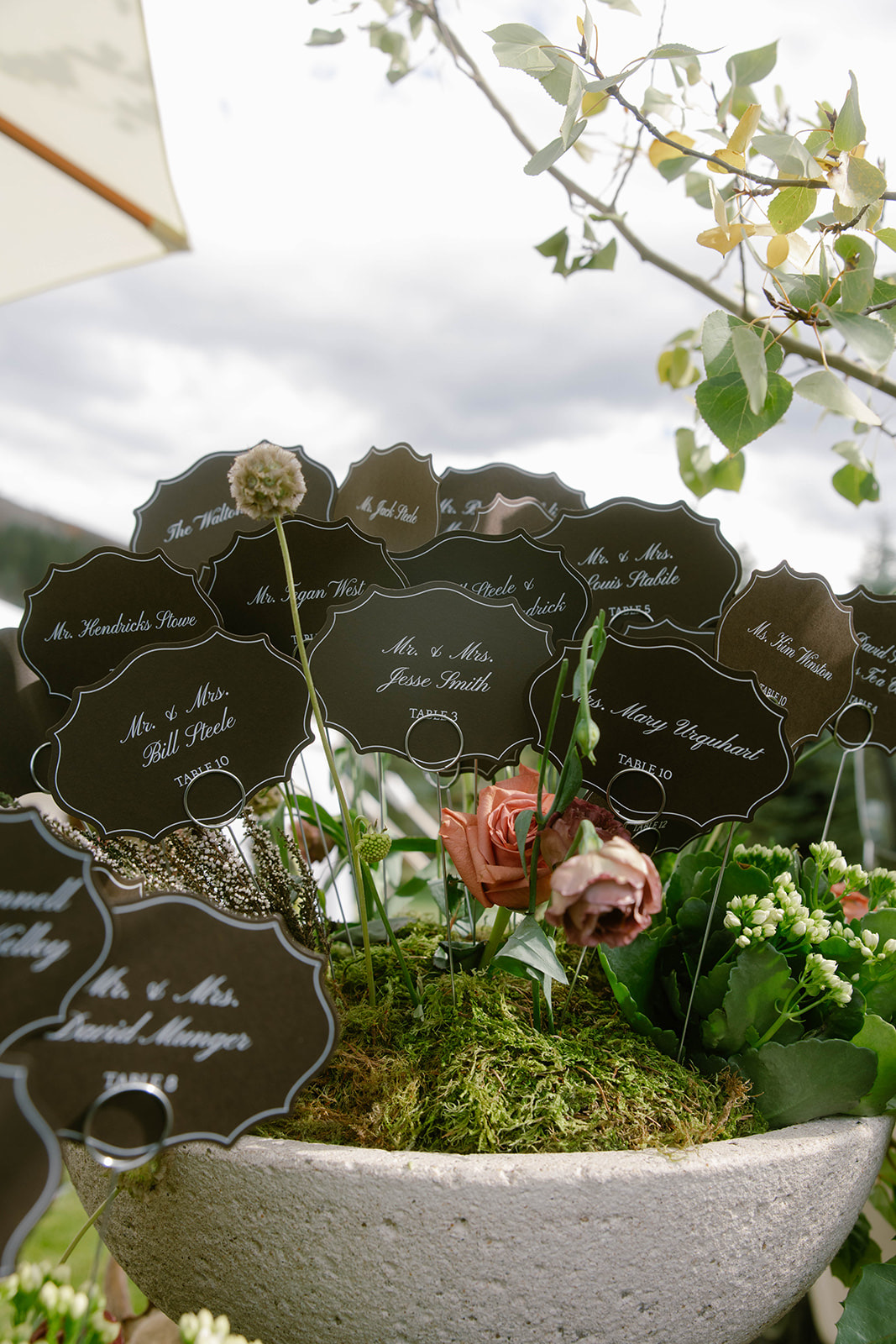 Scalloped die-cut escort cards nestled in a stone urn of greenery and roses.