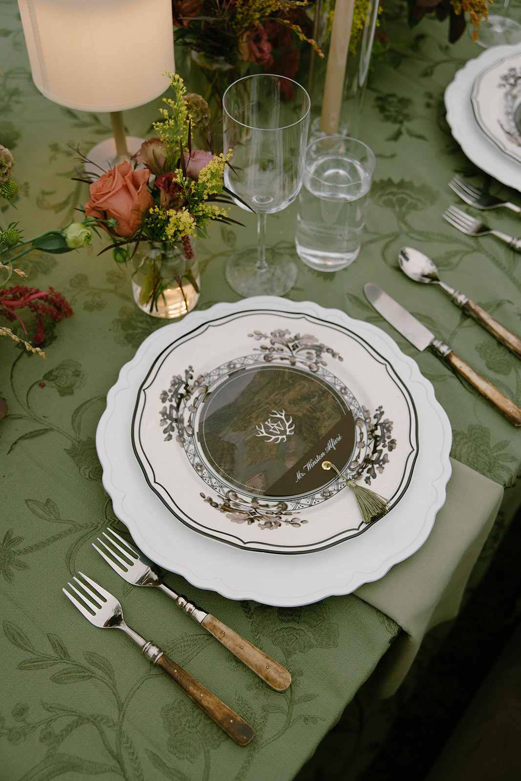 A place setting with glassware and a bud vase of garden roses on sage linen.