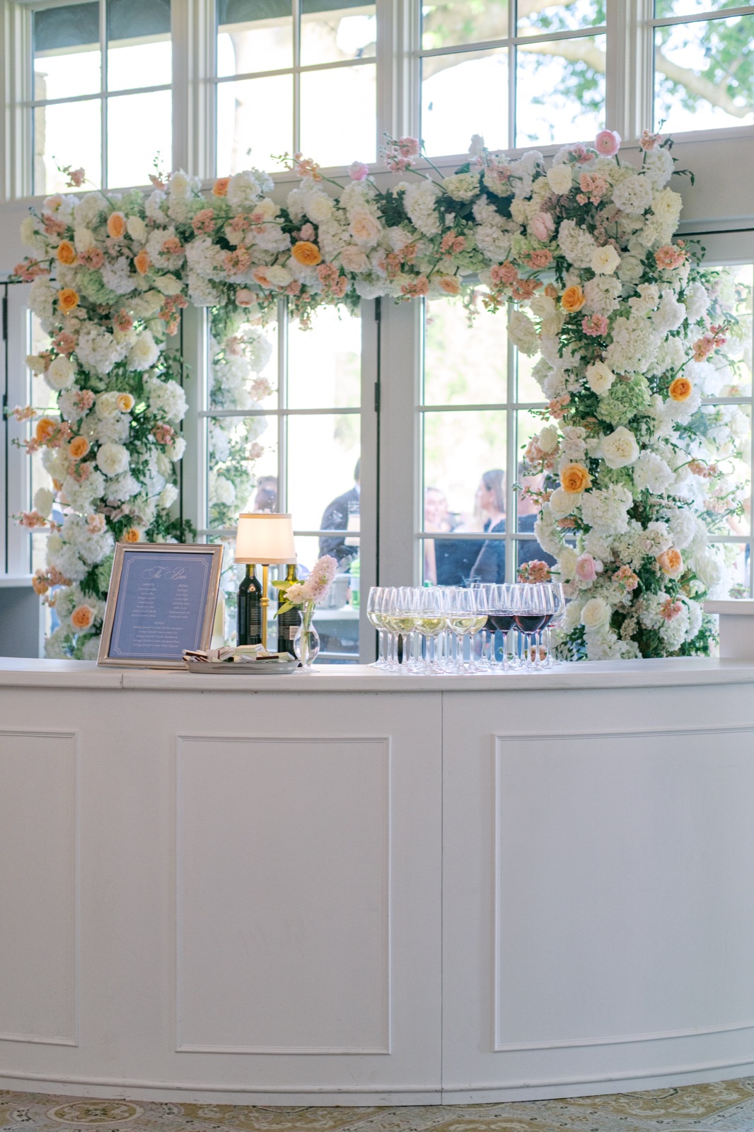A reception bar crowned with a lush arch of white and peach flowers, with a framed menu below.
