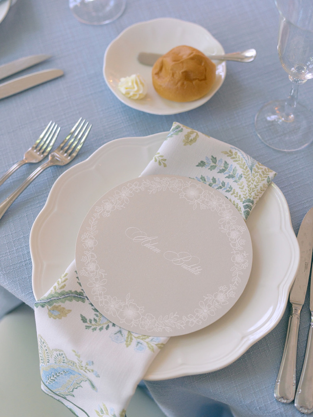 A round place card on a scalloped plate with a fern-printed napkin on soft blue linen.
