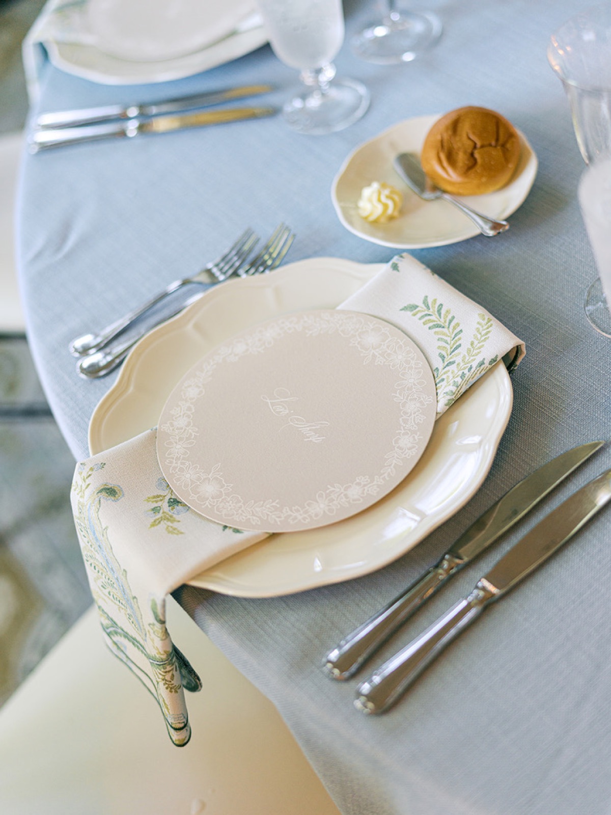 A corner place setting with a round menu, fern napkin, and bread plate on blue linen.