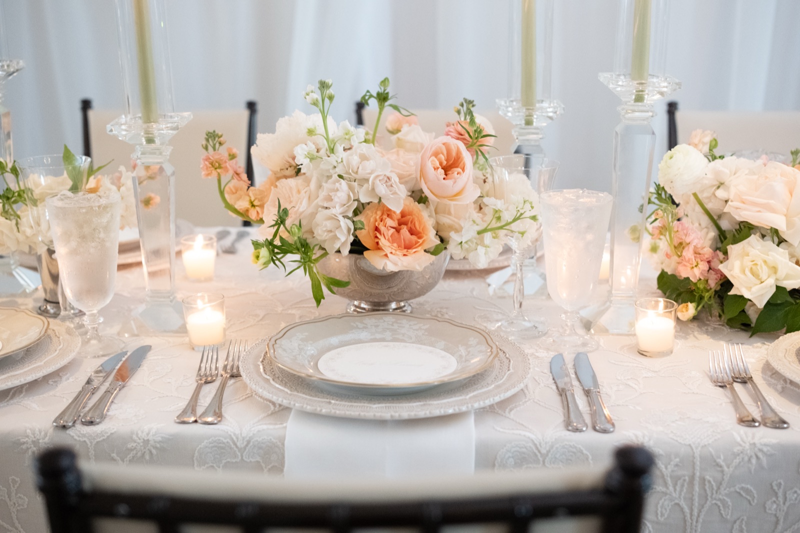 A gold-rimmed place setting with lace charger, monogrammed napkin, and peach florals beyond.