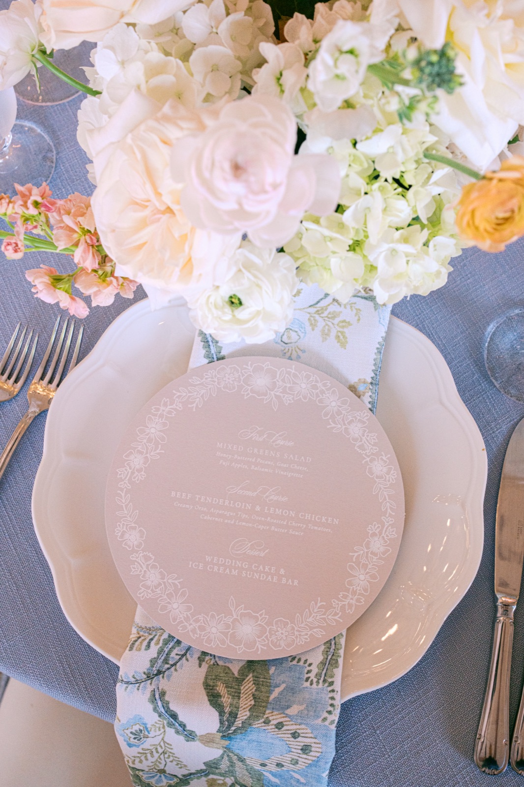 A round wedding menu with a delicate floral border resting on a scalloped plate and soft blue floral napkin.