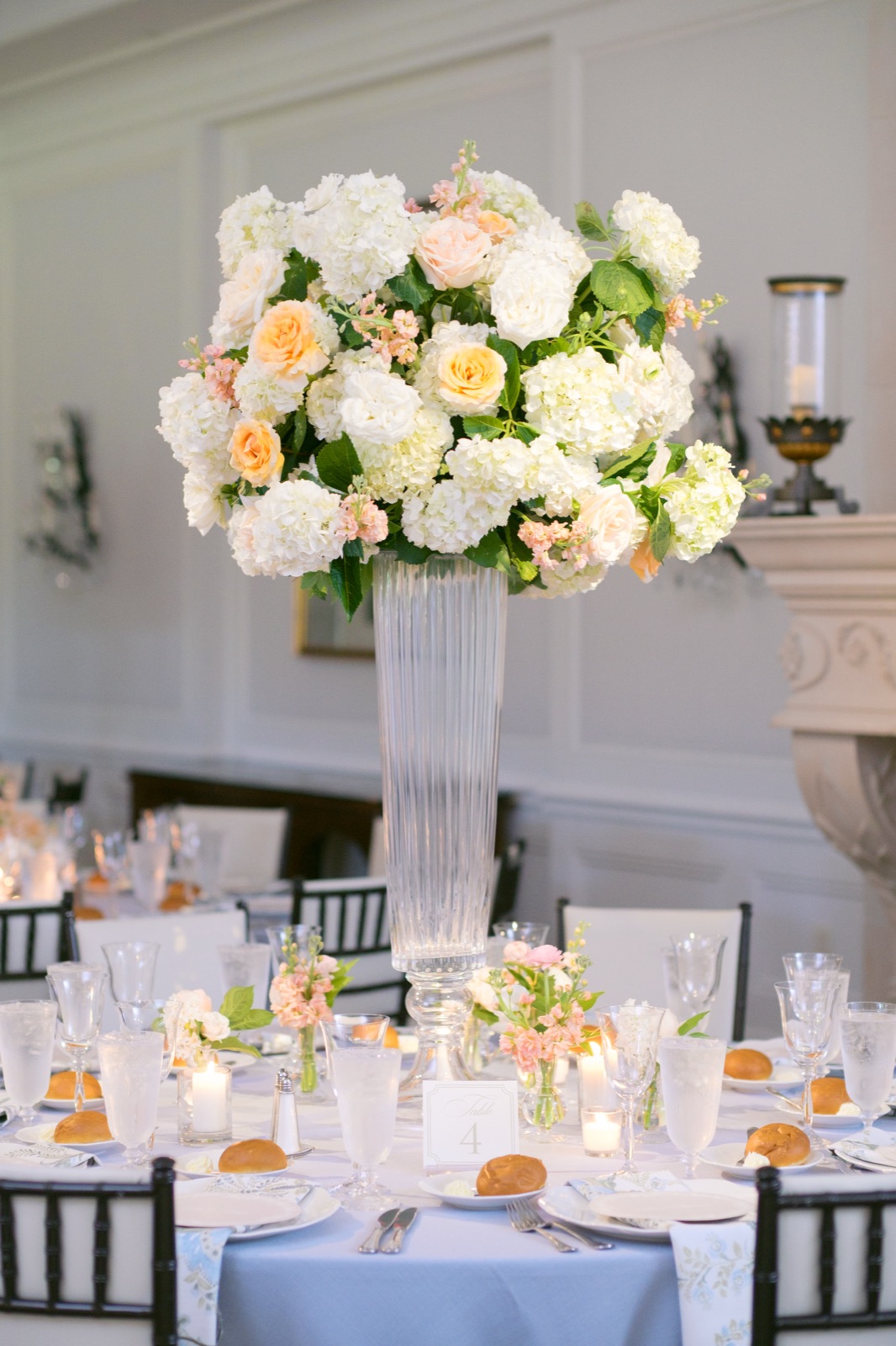 A tall arrangement of garden roses and hydrangea over a reception table set with round menus and soft blue linens.