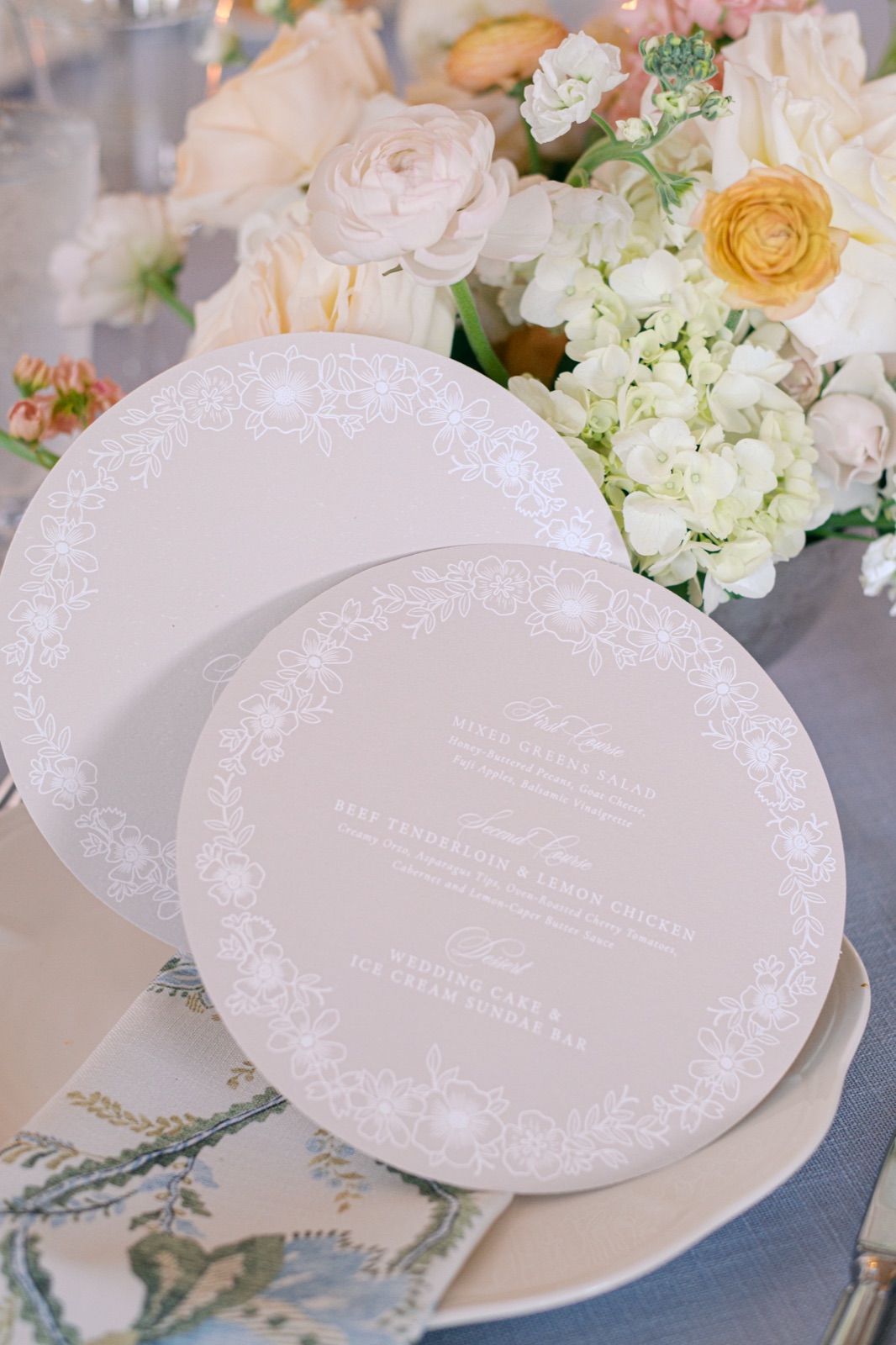 Round wedding menu cards in soft taupe with a delicate white floral border and calligraphy, resting on a place setting.