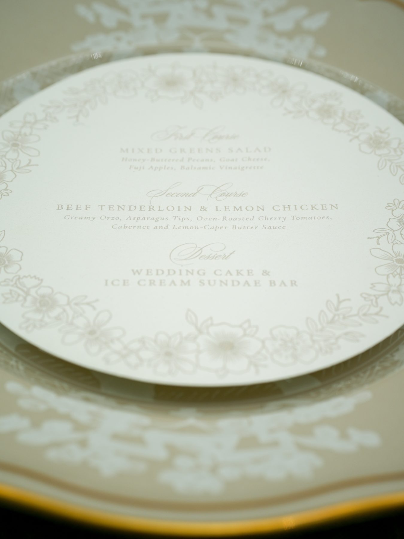 A close-up of a soft beige round wedding menu resting inside a gold-rimmed china charger with a delicate floral border.
