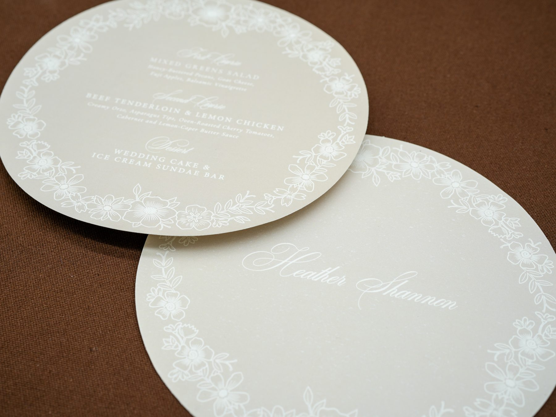 An overhead detail of two layered round wedding menu cards in cream with delicate white floral borders, on dark brown linen.