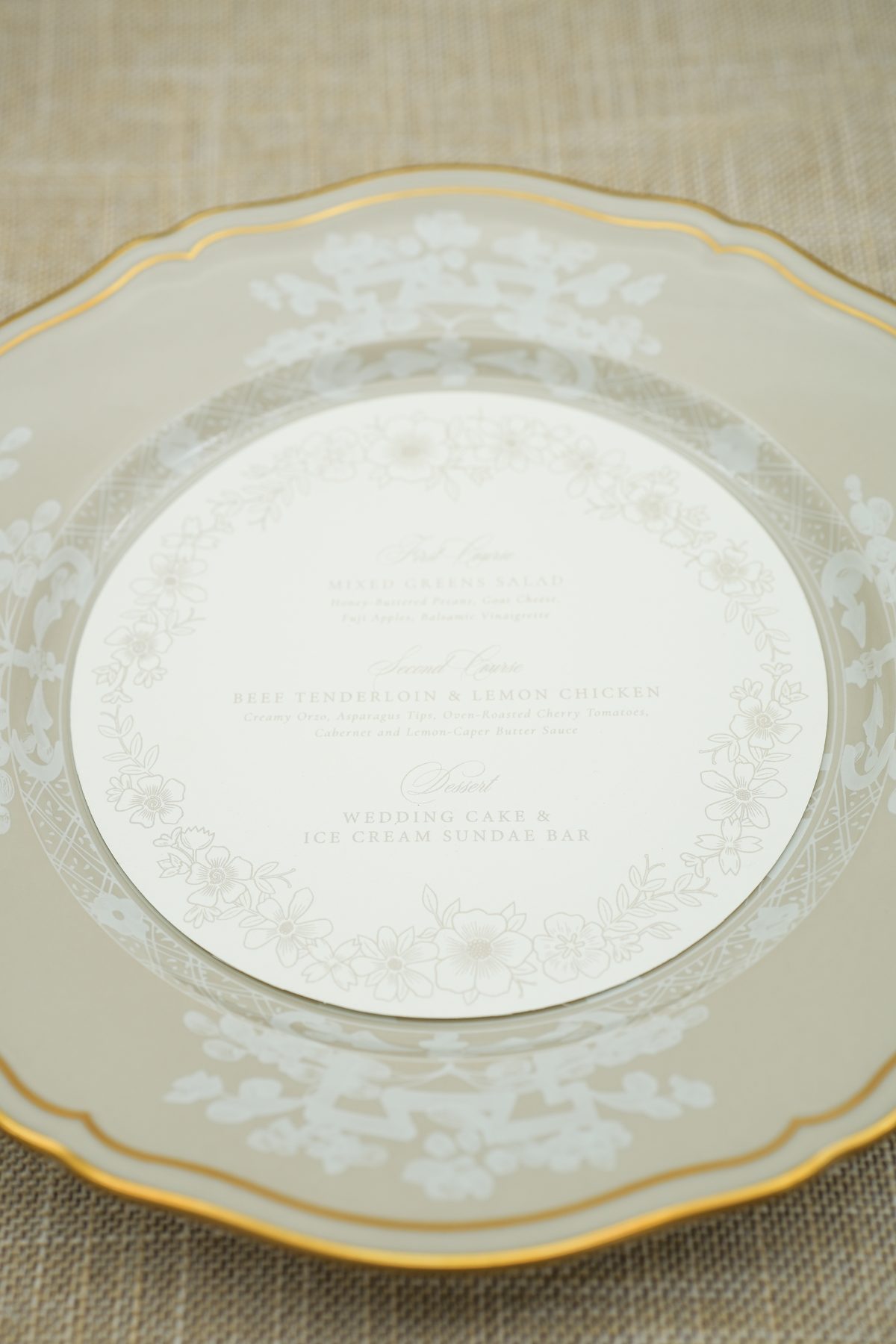 Soft beige round wedding menu card resting on a gold-rimmed china charger with a delicate floral border.