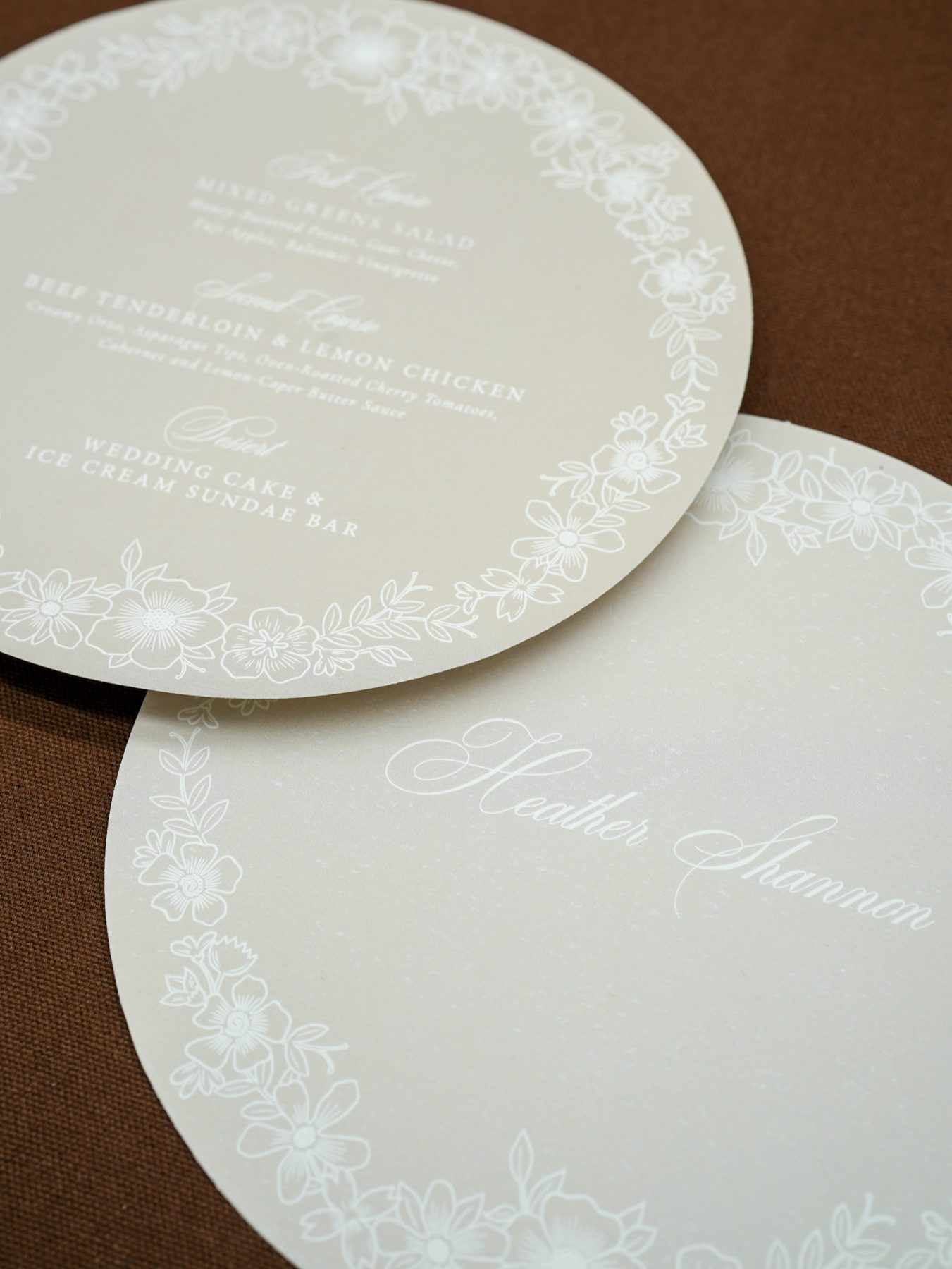 Layered round wedding stationery in soft cream with delicate white floral borders, paired with a script calligraphy place card, by Little Village Creative Co.