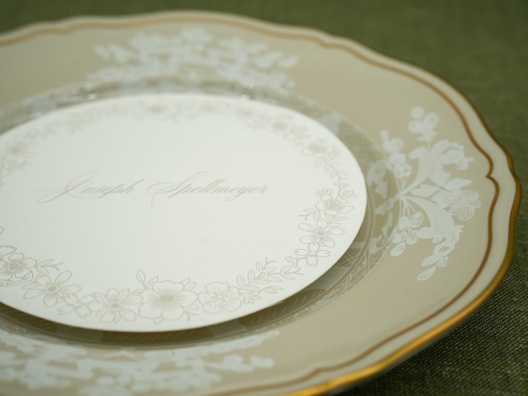 An overhead round wedding place card with delicate floral borders, resting on dark brown linen beside a gold-rimmed china plate.