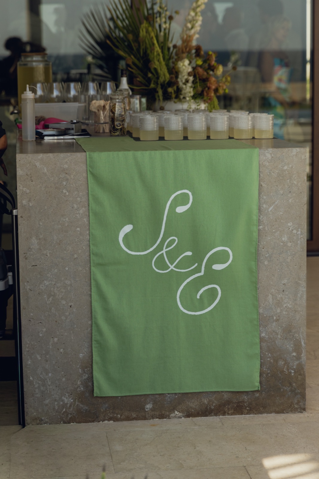 A stone bar draped with a green monogram banner at a beach reception.