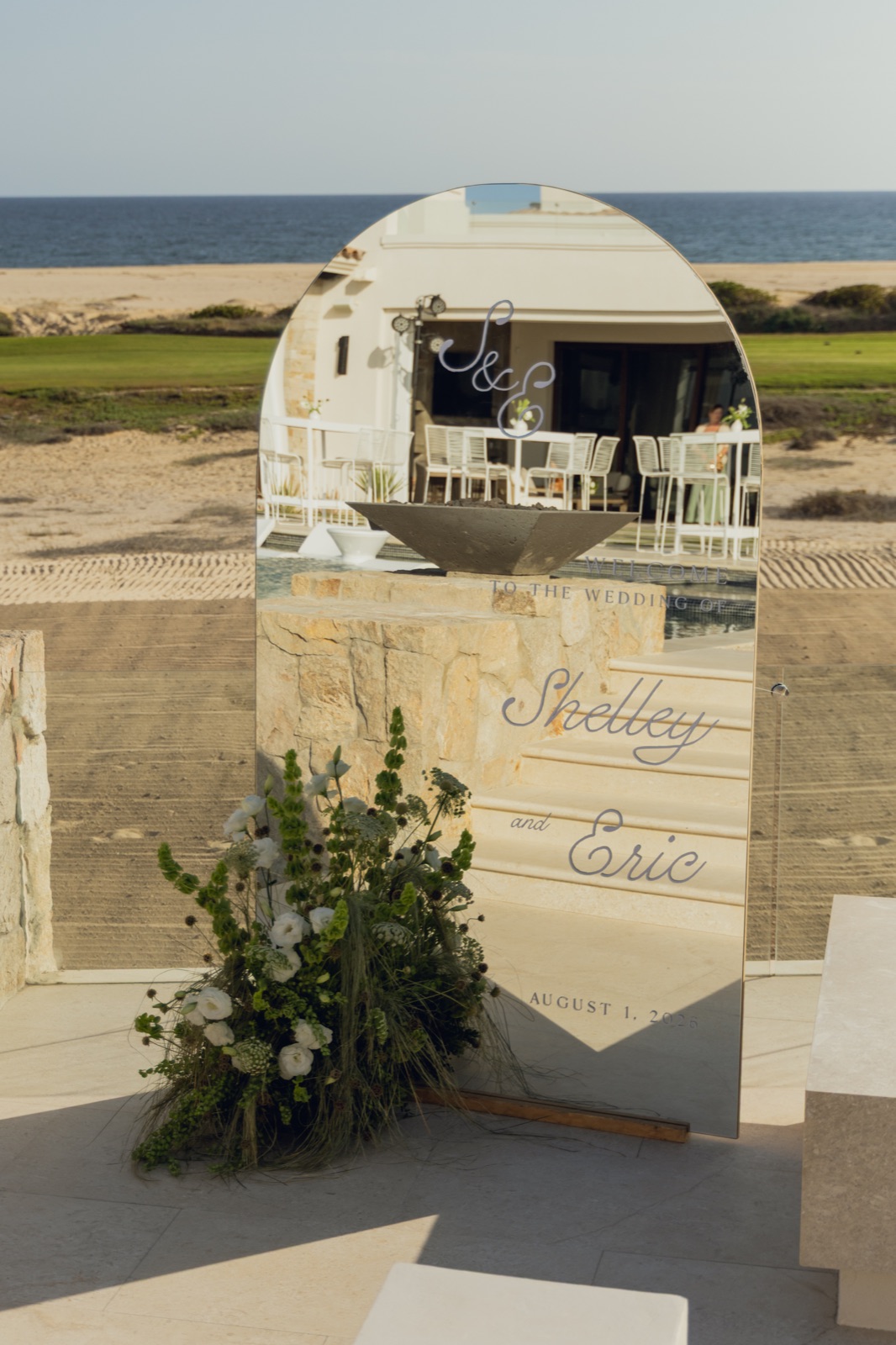 A mirrored welcome sign in script, styled with greenery on the sand.