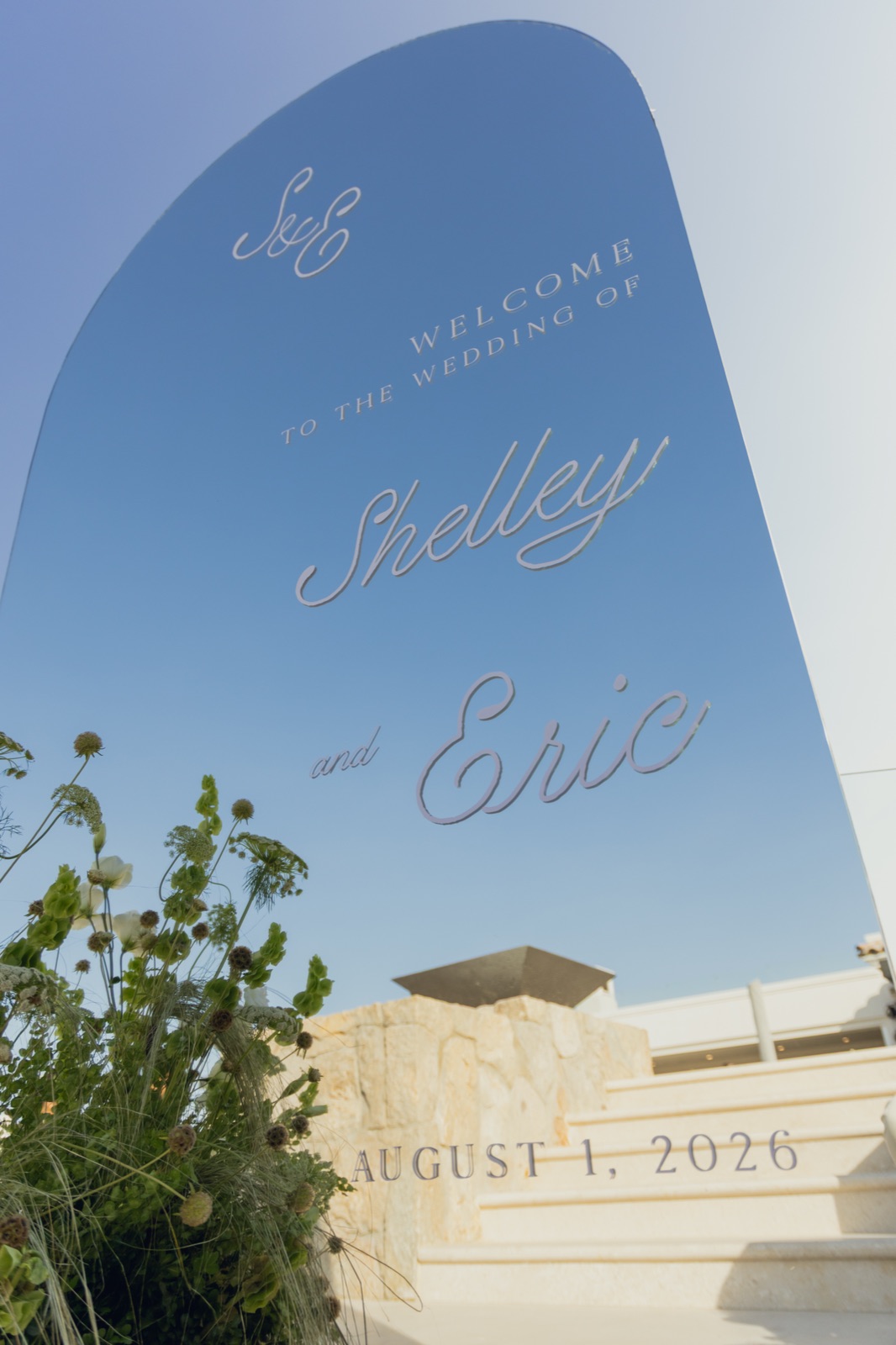 A mirrored arch welcome sign lettered for the wedding day against the sky.