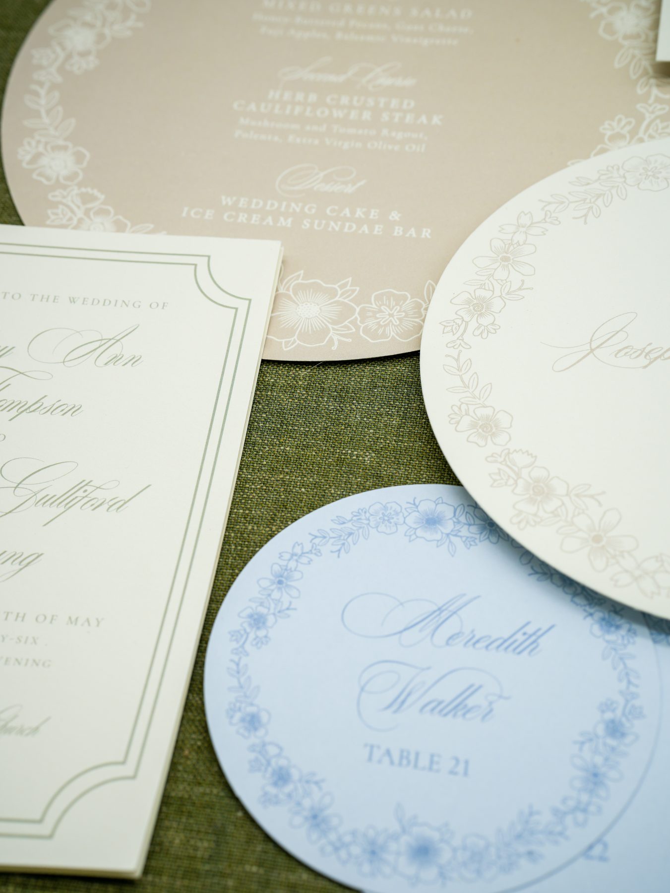 A fanned spring wedding suite of letterpress invitation, round menus, and round blue floral place cards on olive linen.