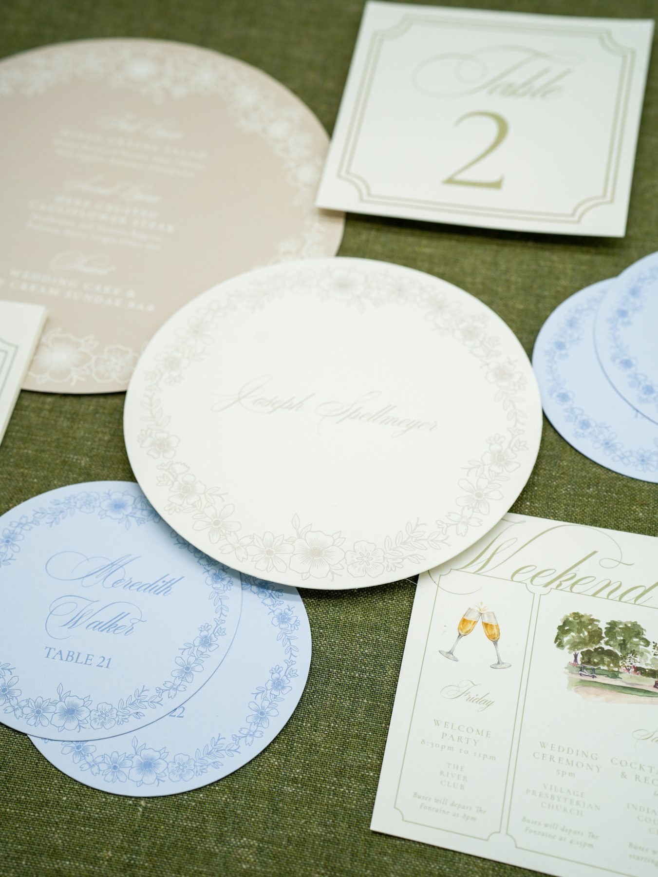 A garden wedding day-of arrangement of a round menu, soft blue round place cards, a framed table number, and a weekend itinerary on olive linen.