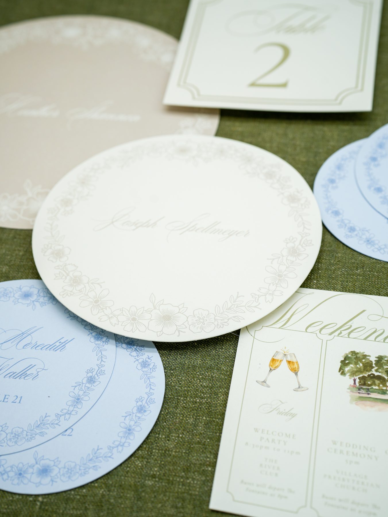 An arrangement of round menus, soft blue round place cards, and a framed table number on olive linen.