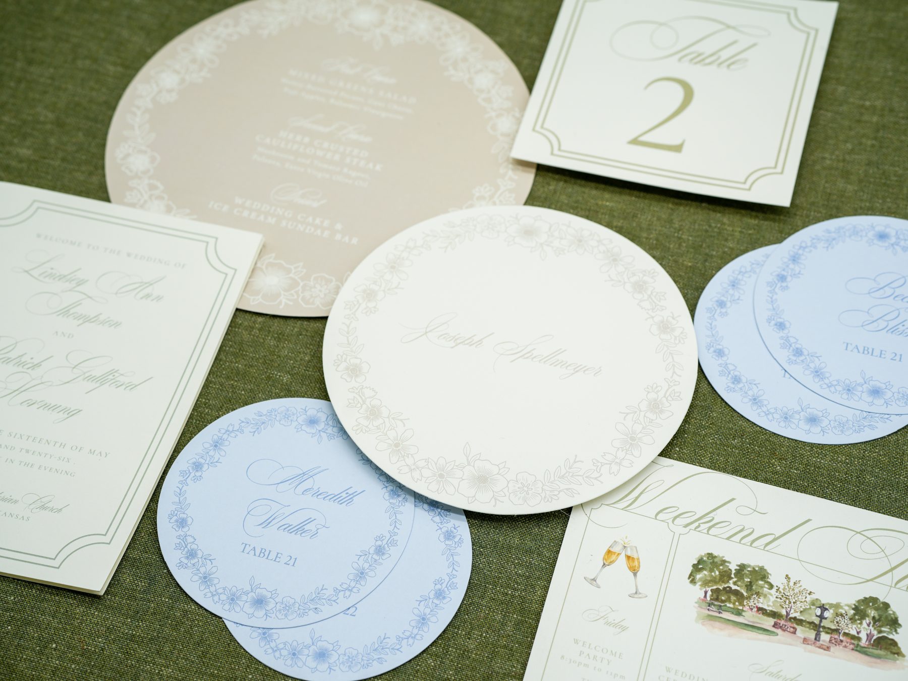An overhead view of a spring wedding place setting with round menu, blue round place cards, table number, and a charger plate on olive linen.
