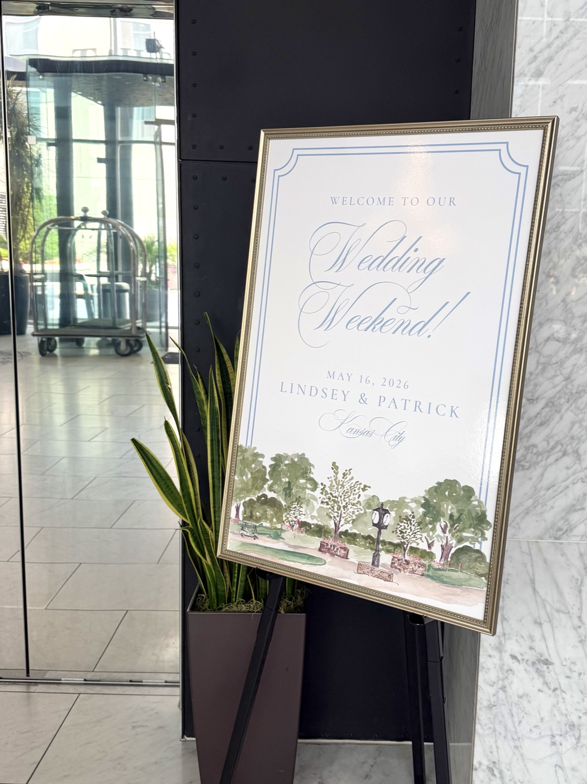 A gold-framed wedding welcome sign on an easel featuring soft blue calligraphy and a watercolor park illustration.